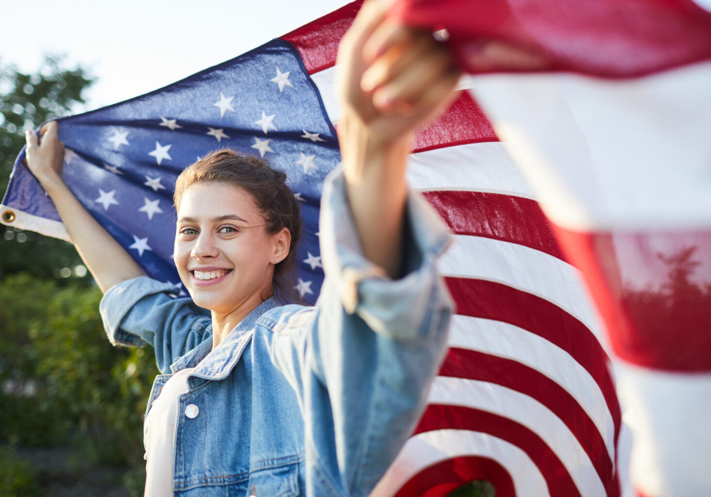 Portrait of positive beautiful teenage girl in denim jacket inspired with American culture raising American flag above head